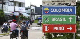 foto mostra duas pessoas andando de moto pela rua, sem uso do capacete, uma ao lado da outra. Em primeiro plano, há uma placa de localização, com os nomes Colombia, Brasil e Peru. Esses países fazem parte da integração sul-americana