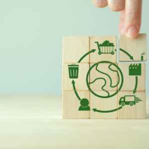 Produção de insumos virgens cresce e ameaça avanço da economia circular Blocos de madeira com ícones que representam a circularidade de um produto, como produção, consumo, descarte e reutilização, com a mão posicionando o bloco que representa a etapa de descarte