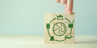 Blocos de madeira com ícones que representam a circularidade de um produto, como produção, consumo, descarte e reutilização, com a mão posicionando o bloco que representa a etapa de descarte