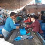 Catadores trabalhando em uma linha de produção de reciclagem, separando materiais em um galpão com pilhas de lixo ao fundo.