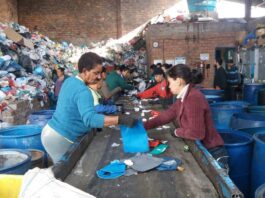 Catadores trabalhando em uma linha de produção de reciclagem, separando materiais em um galpão com pilhas de lixo ao fundo.
