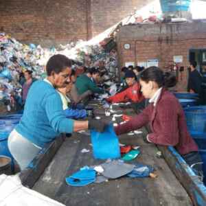 Governo revoga decreto de importação de resíduos recicláveis após mobilização de catadores Catadores trabalhando em uma linha de produção de reciclagem, separando materiais em um galpão com pilhas de lixo ao fundo.