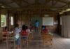 Sala de aula simples em madeira com alunos e professor, mostrando a necessidade de acesso à tecnologia em comunidades ribeirinhas.