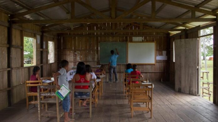 Sala de aula simples em madeira com alunos e professor, mostrando a necessidade de acesso à tecnologia em comunidades ribeirinhas.