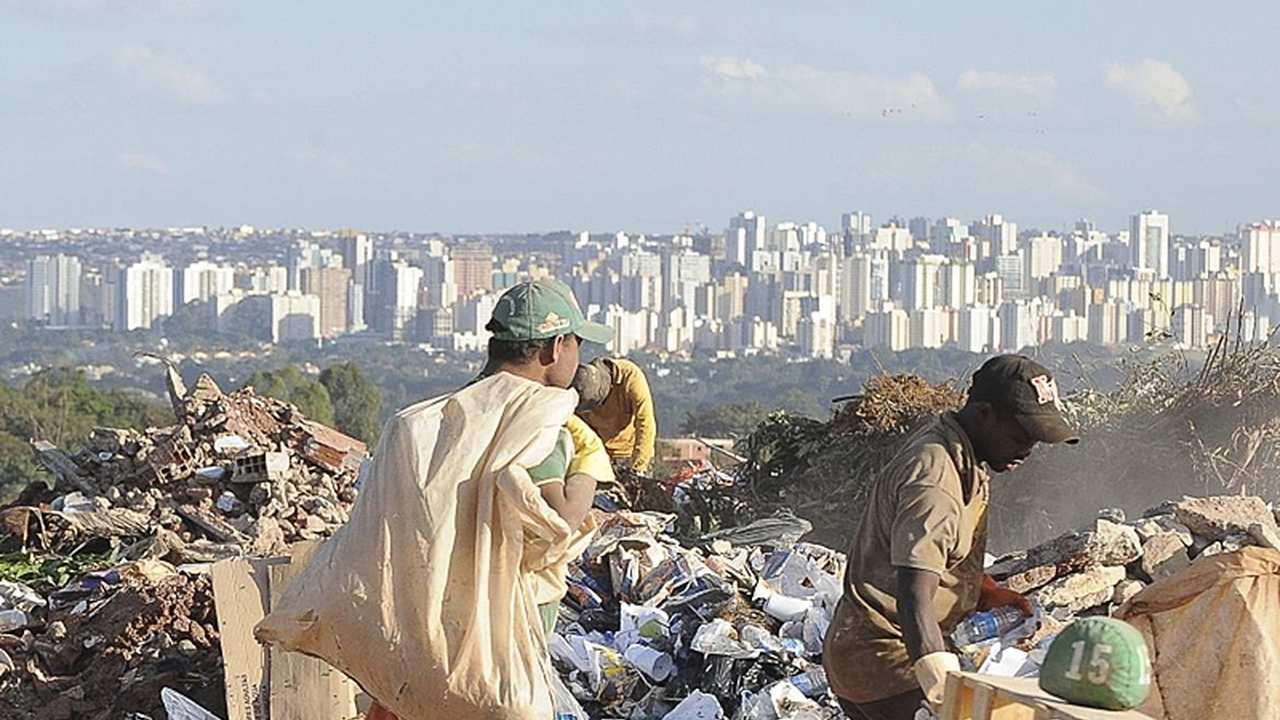 Trabalhadores catando lixo em um lixão, com uma cidade ao fundo.