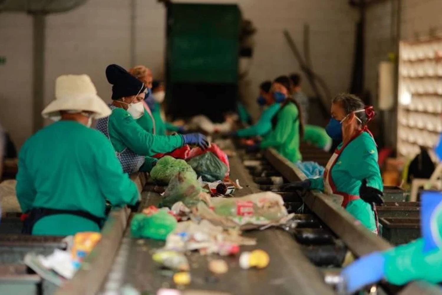 A virada verde do Brasil: Decreto federal obriga uso de plástico reciclado em embalagens 2 Trabalhadores em uma linha de montagem de reciclagem, usando máscaras e luvas, classificam e separam lixo reciclável.