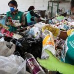 Três mulheres trabalhando na coleta de lixo reciclável, vestindo uniformes e luvas, em frente a um monte de garrafas e sacolas plásticas.