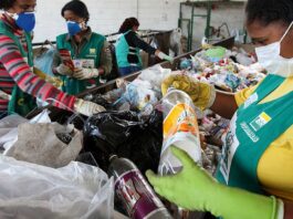 Três mulheres trabalhando na coleta de lixo reciclável, vestindo uniformes e luvas, em frente a um monte de garrafas e sacolas plásticas.
