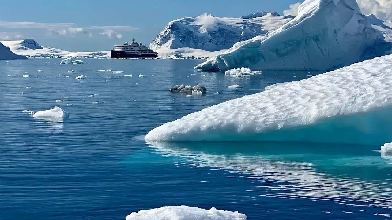 Um cruzeiro navega em águas azuis geladas da Antártida, com icebergs e montanhas nevadas ao fundo.