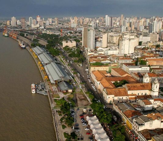 Vista aérea de uma cidade litorânea com edifícios, um rio e um cais com barracas brancas e barcos atracados.