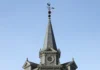 Ninho de vespa-asiática descoberto a 55 metros de altura no pináculo da igreja: “desafio de exterminá-los”, na Bélgica Torre afilada de igreja em enquadramento central 16:9, com ninho escuro de vespa posicionado imediatamente sob o cata-vento no ponto mais alto, céu limpo e sem marcações gráficas.