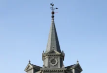 Ninho de vespa-asiática descoberto a 55 metros de altura no pináculo da igreja: “desafio de exterminá-los”, na Bélgica Torre afilada de igreja em enquadramento central 16:9, com ninho escuro de vespa posicionado imediatamente sob o cata-vento no ponto mais alto, céu limpo e sem marcações gráficas.