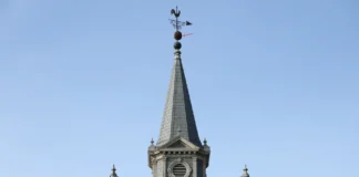 Ninho de vespa-asiática descoberto a 55 metros de altura no pináculo da igreja: “desafio de exterminá-los”, na Bélgica Torre afilada de igreja em enquadramento central 16:9, com ninho escuro de vespa posicionado imediatamente sob o cata-vento no ponto mais alto, céu limpo e sem marcações gráficas.