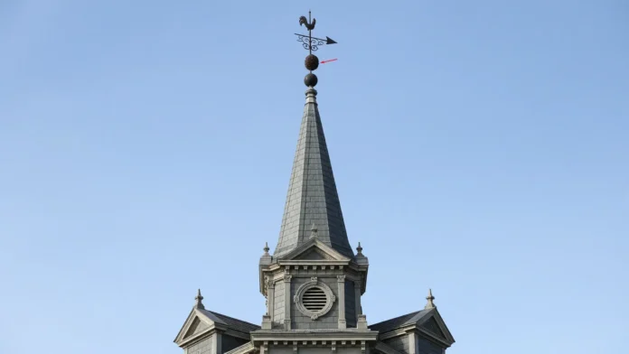 ninho-vespa-asiatica-pinaculo-igreja-belgica Torre afilada de igreja em enquadramento central 16:9, com ninho escuro de vespa posicionado imediatamente sob o cata-vento no ponto mais alto, céu limpo e sem marcações gráficas.