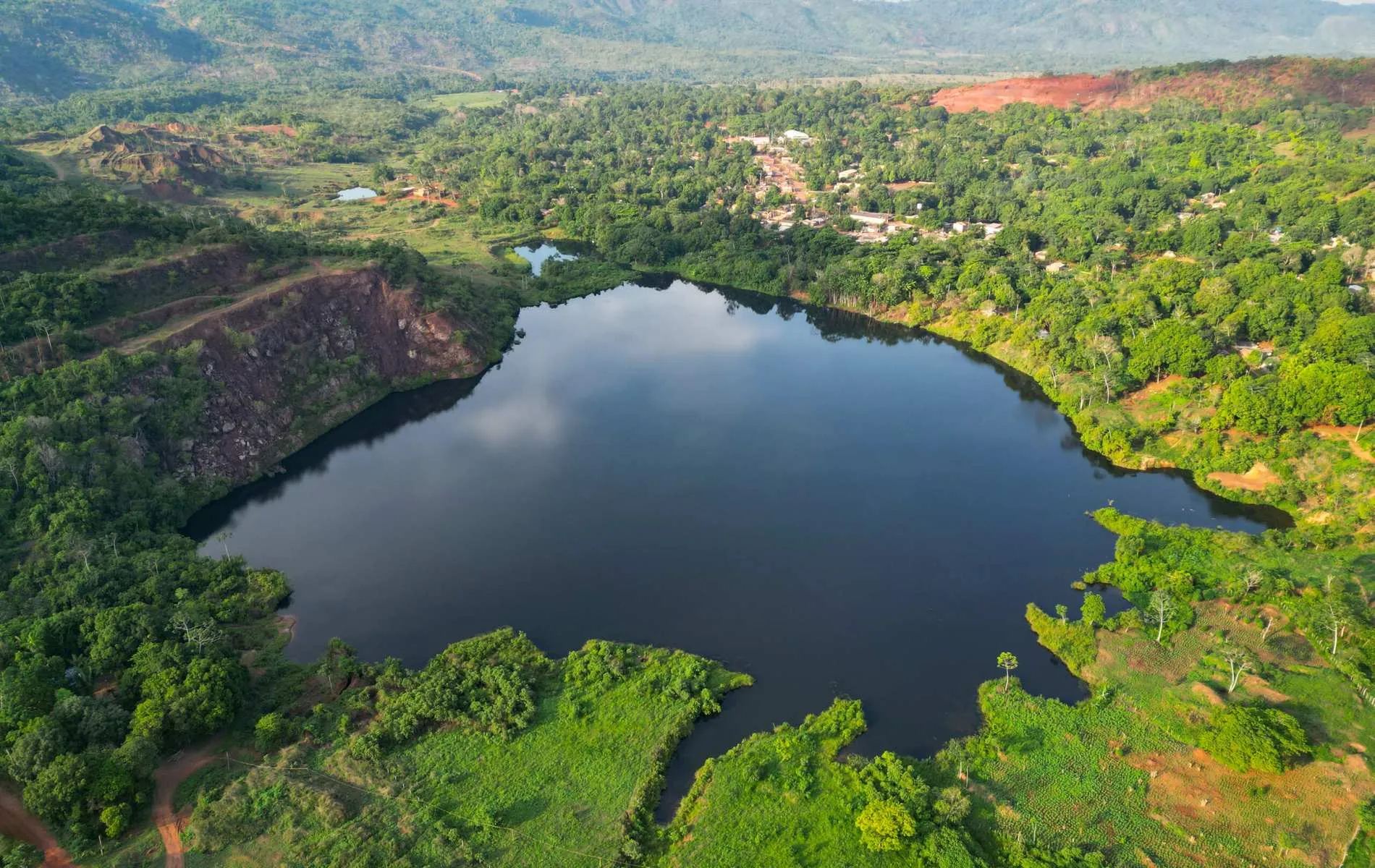 Lago azul profundo cercado por vegetação verde em Serra Pelada, com uma vila no topo da colina ao fundo