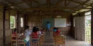 Sala de aula simples em madeira com alunos e professor, mostrando a necessidade de acesso à tecnologia em comunidades ribeirinhas.