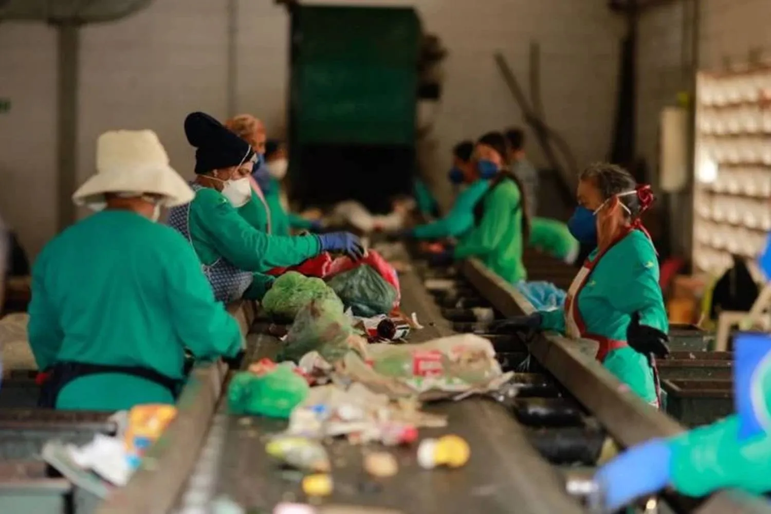 Trabalhadores em uma linha de montagem de reciclagem, usando máscaras e luvas, classificam e separam lixo reciclável.