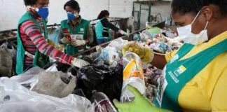 Três mulheres trabalhando na coleta de lixo reciclável, vestindo uniformes e luvas, em frente a um monte de garrafas e sacolas plásticas.