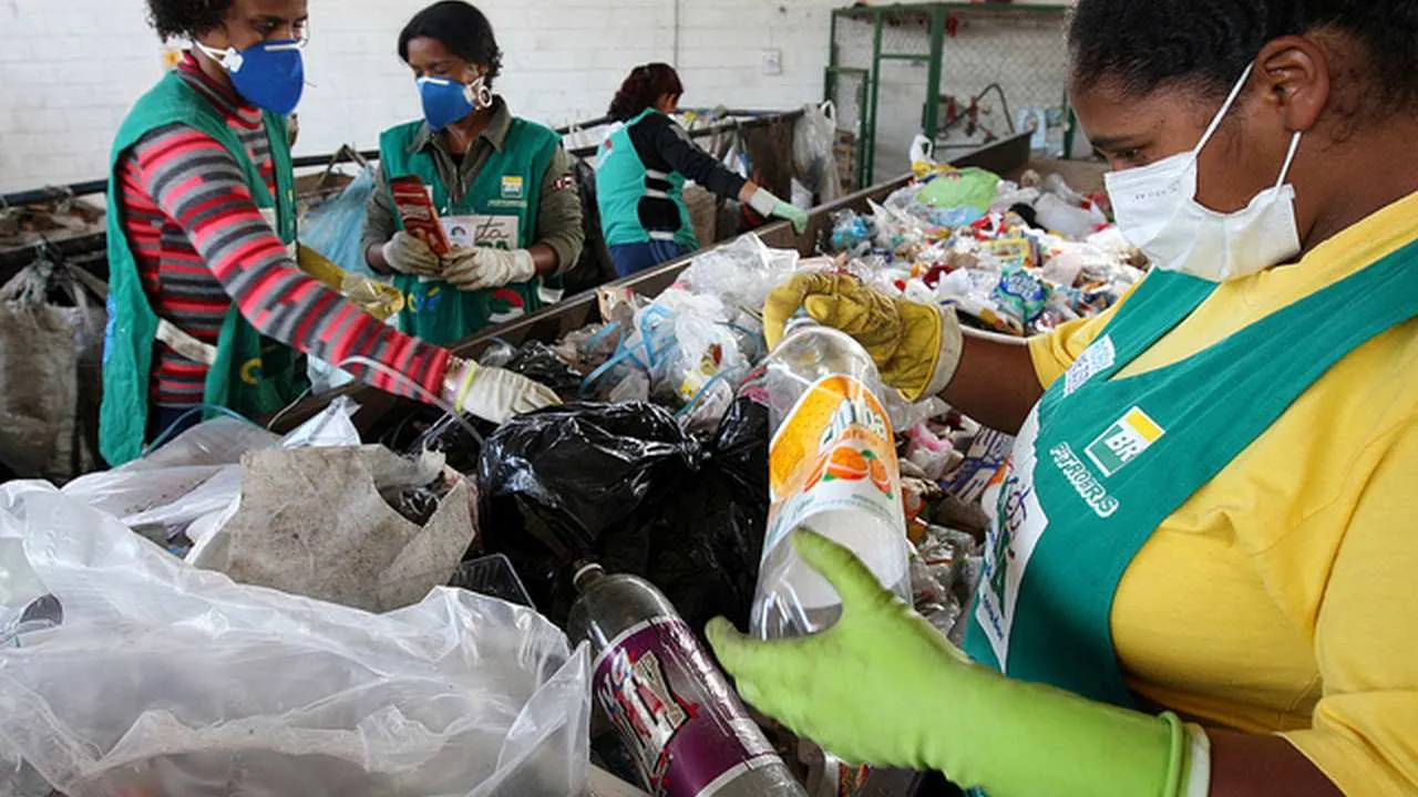 Três mulheres trabalhando na coleta de lixo reciclável, vestindo uniformes e luvas, em frente a um monte de garrafas e sacolas plásticas.