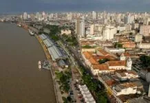 COP 30: ATOS E FATOS Vista aérea de uma cidade litorânea com edifícios, um rio e um cais com barracas brancas e barcos atracados.