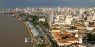 Vista aérea de uma cidade litorânea com edifícios, um rio e um cais com barracas brancas e barcos atracados.